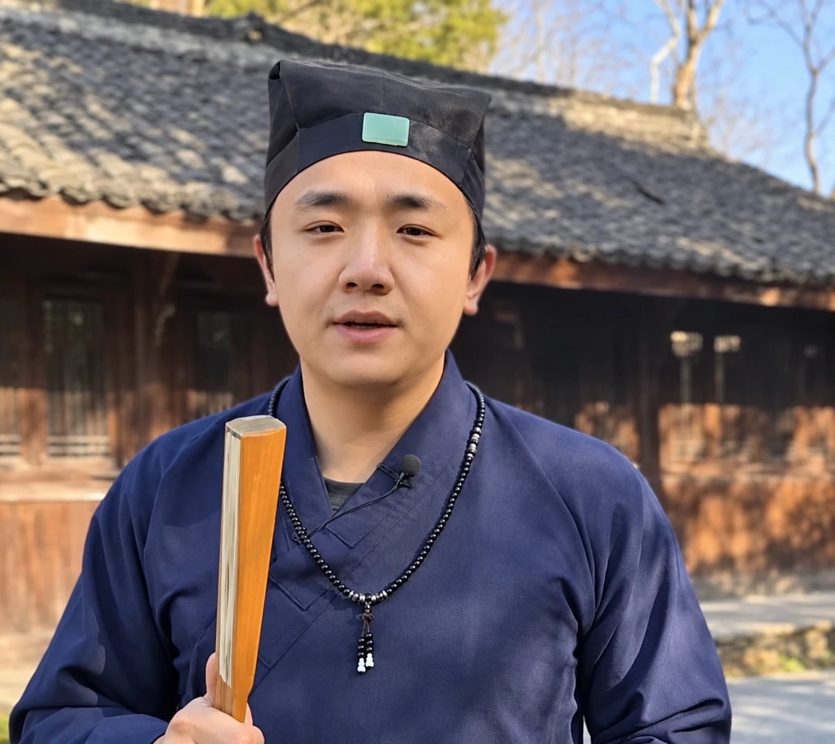 Mike Wang in a navy Daoist robe holding a folded fan outdoors; AI-assisted enhancement of his photograph.